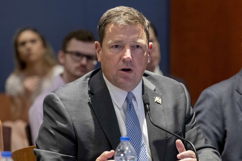 White man with brown hair shown from the chest up wearing a light suit and blue tie while seated and speaking in front of a small microphone.
